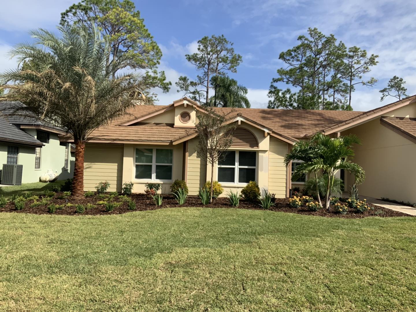 Beige house with brown roof, green lawn, and landscaping under a blue sky.