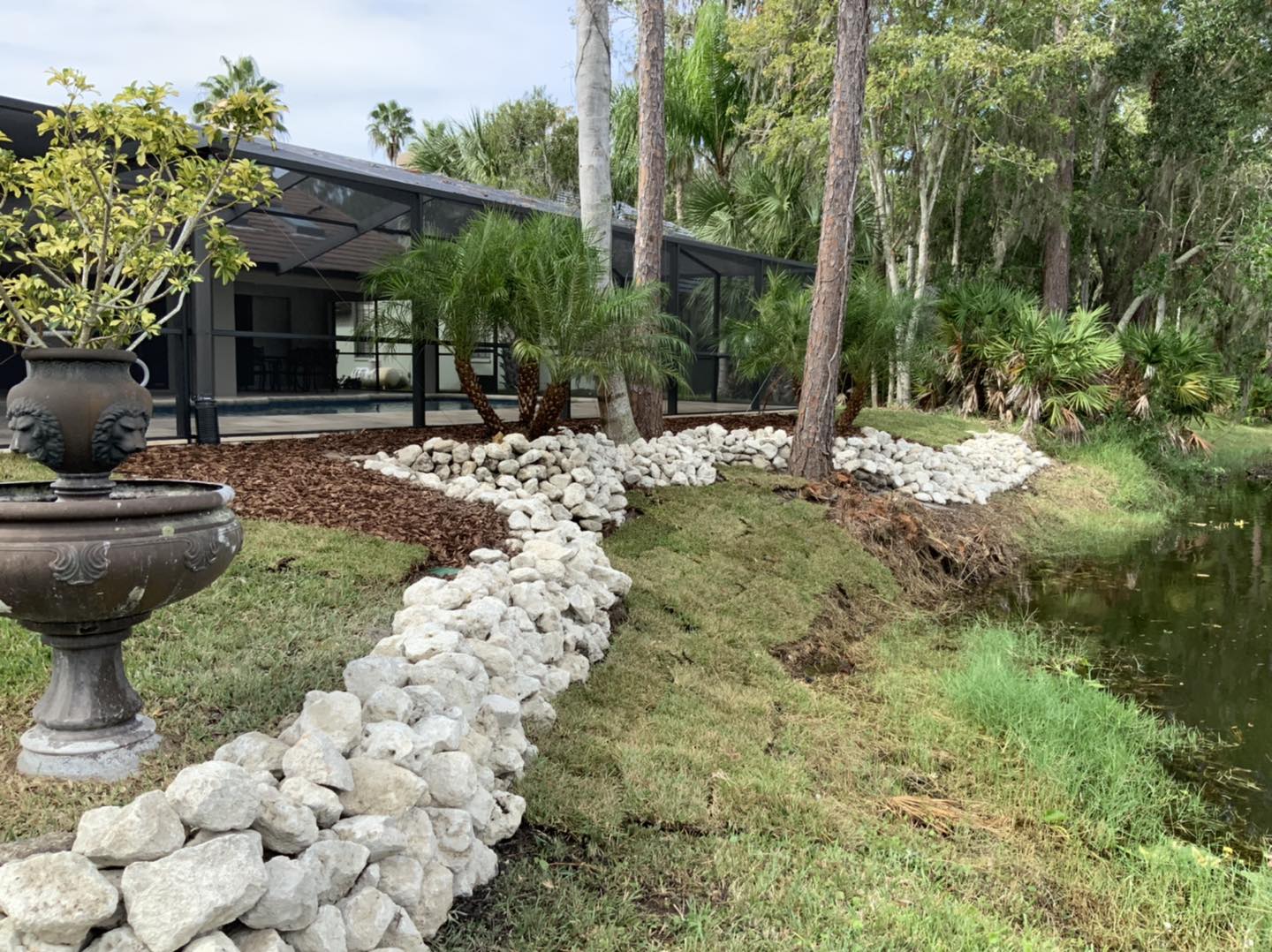 Stone-lined landscaping beside a pond with a house in the background, trees, and a fountain.
