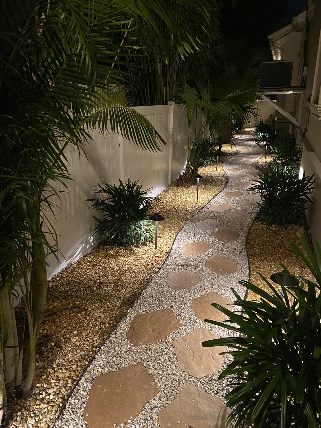A pathway with large stone pavers flanked by gravel, lit by ground lights and plants.