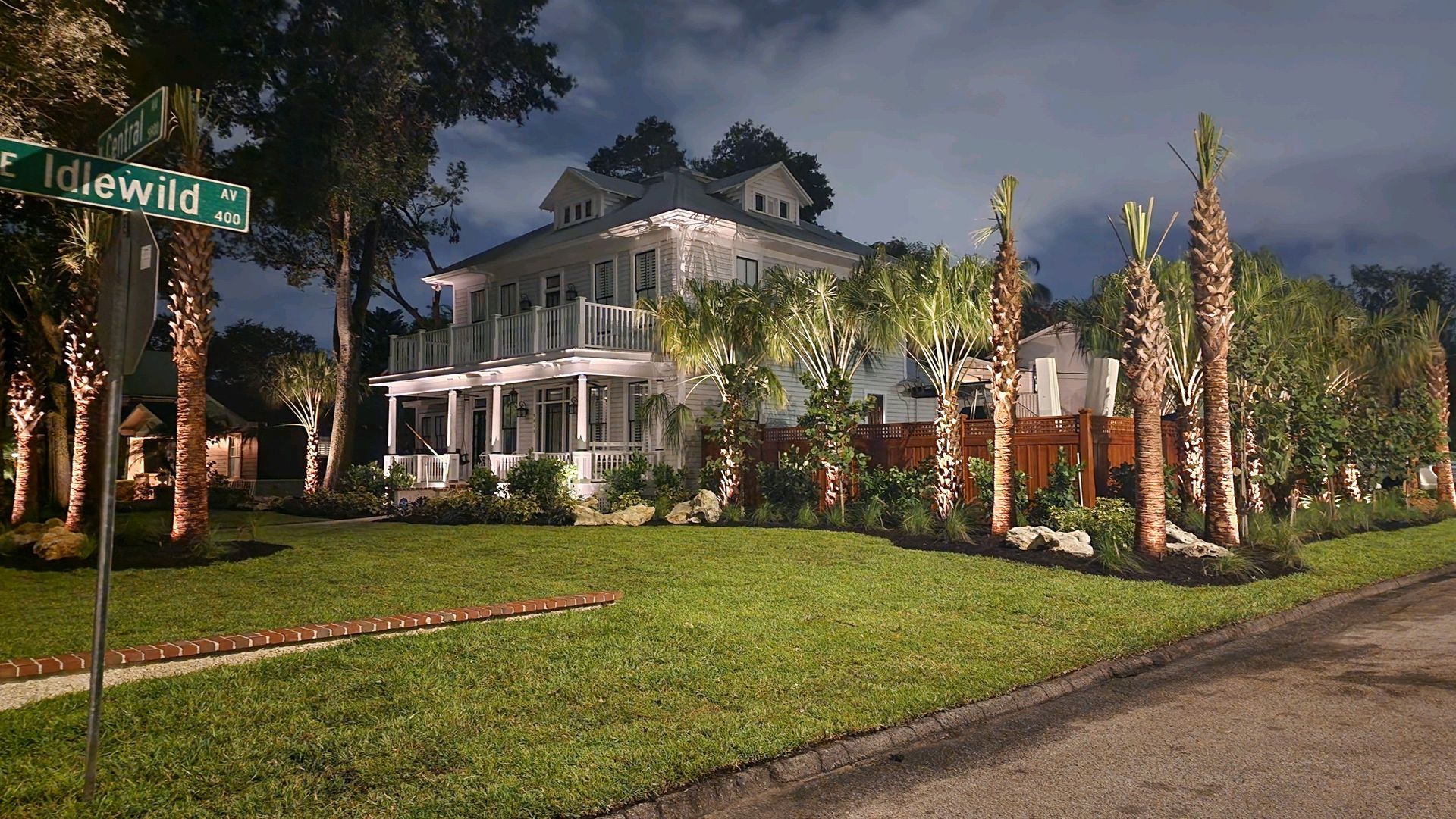 Large white house with multiple stories, surrounded by palm trees and manicured lawn, lit at night.