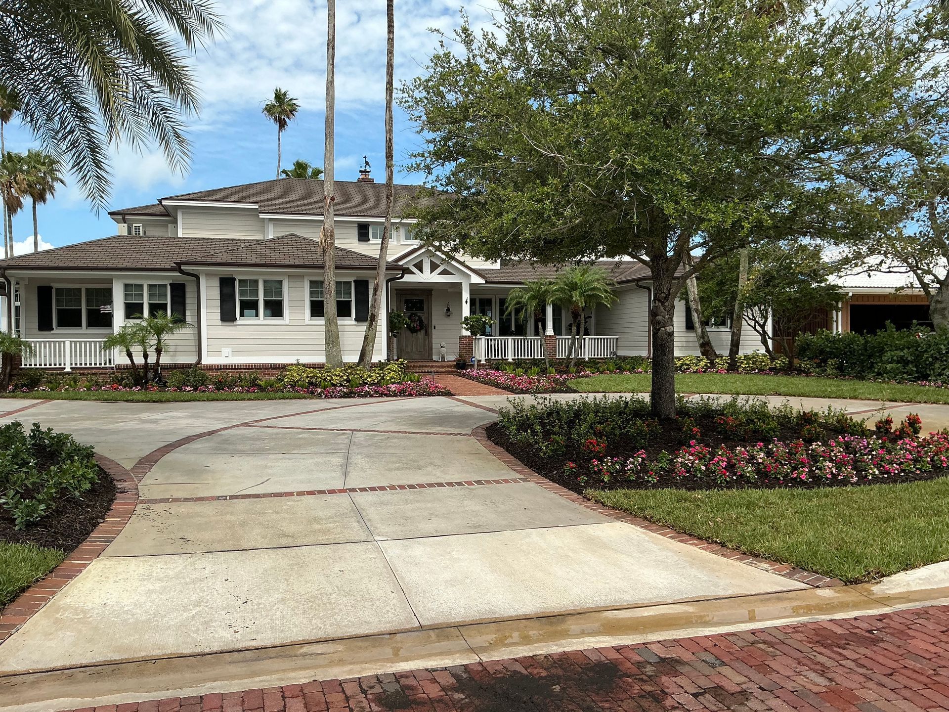Large light-colored house with brick driveway and landscaped flower beds, blue sky.