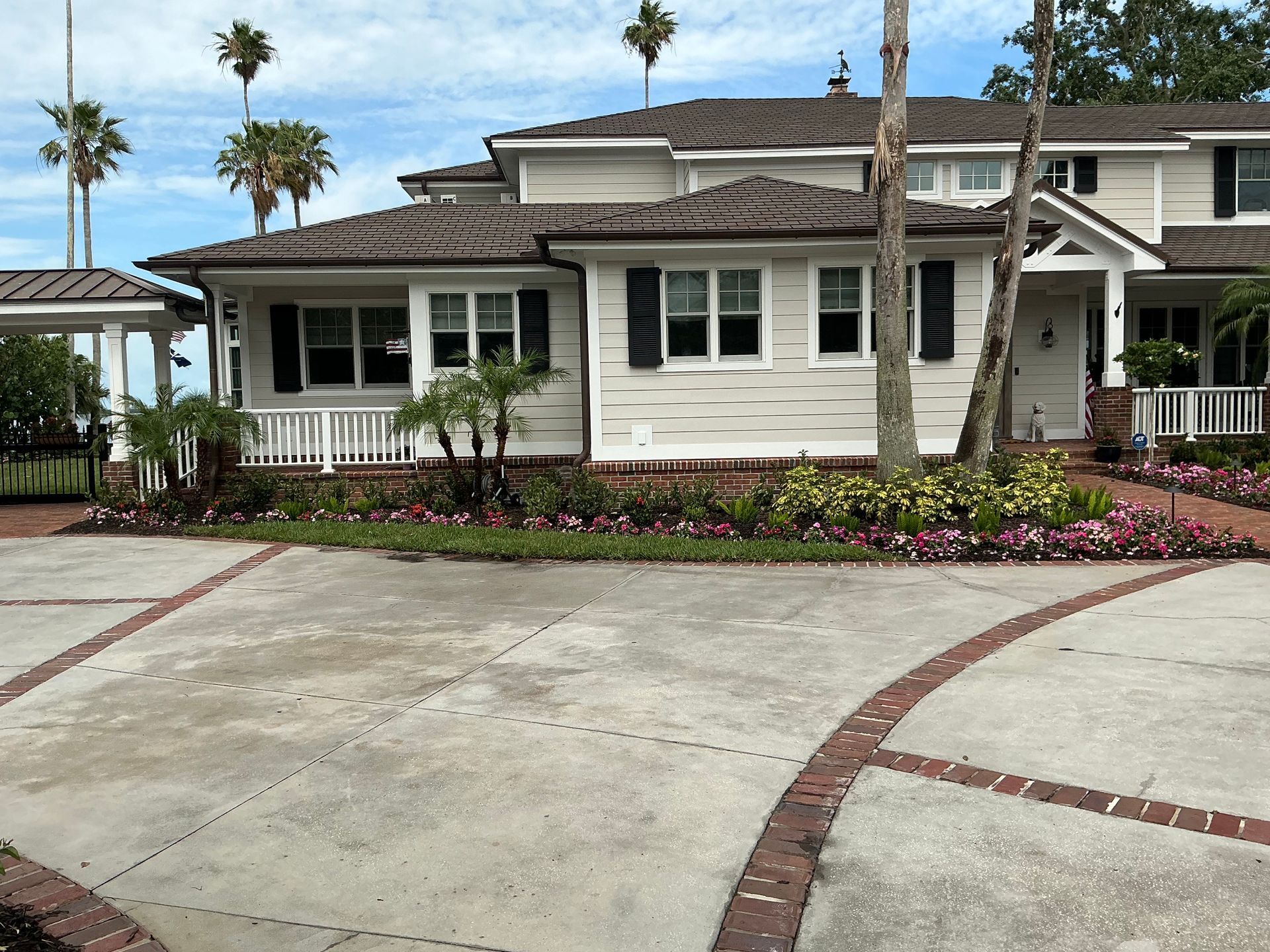 House with white siding, black shutters, and brick driveway, set against a backdrop of palm trees and blue sky.