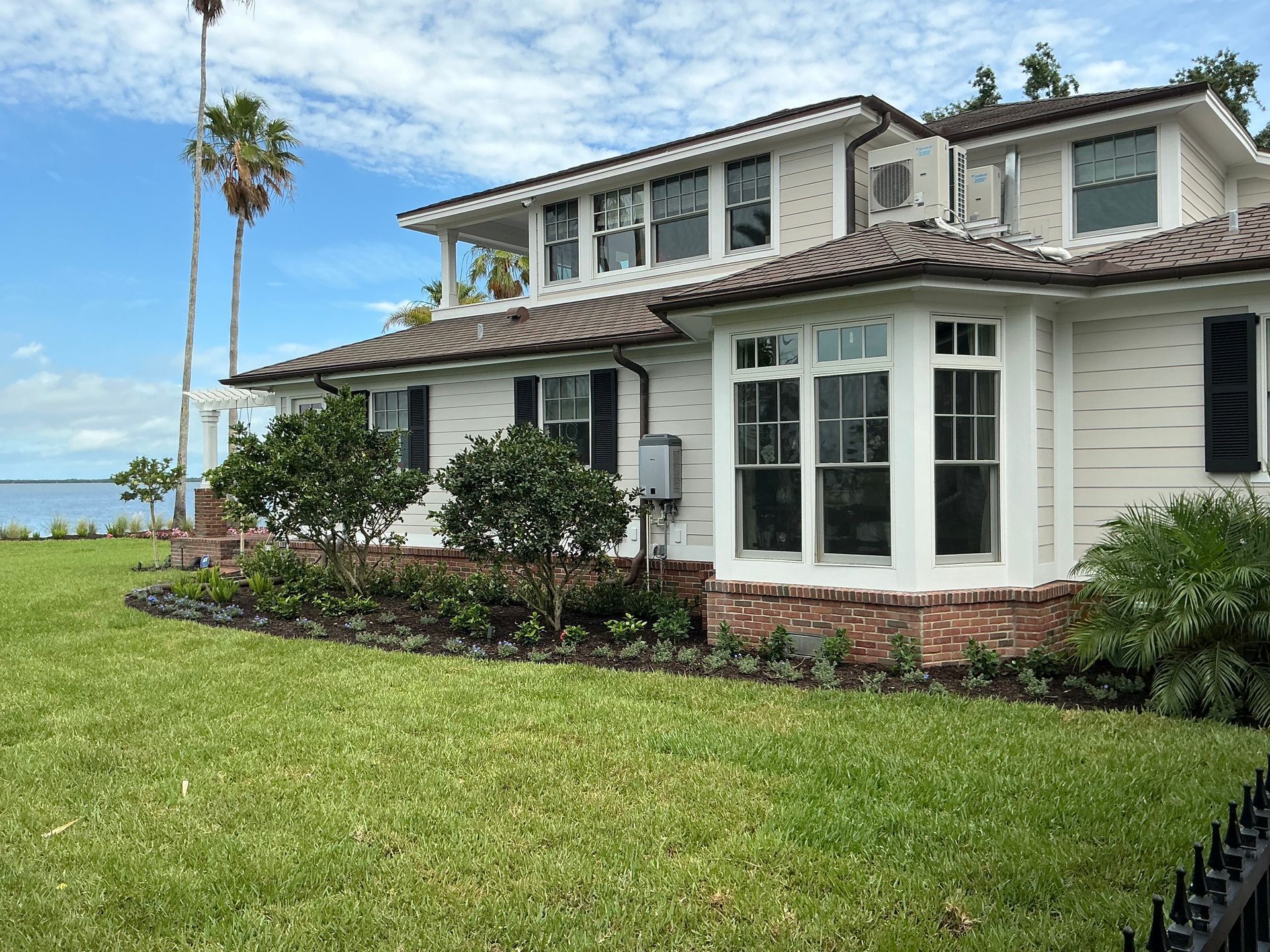 White house with bay window, brick accents, green lawn, and palm trees. Blue sky and water in the background.
