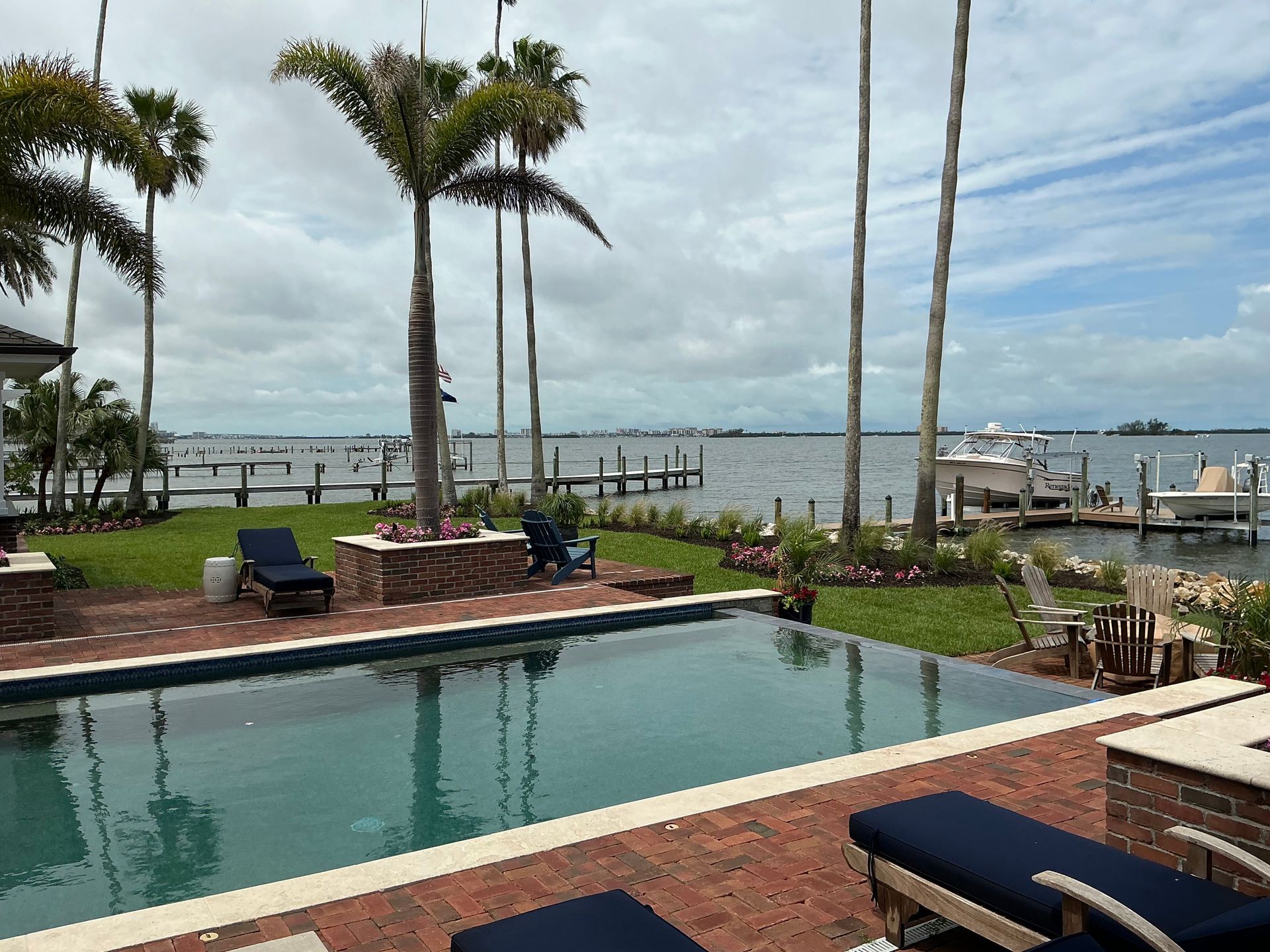 Pool overlooking a waterfront with palm trees, docks, boats, and a cloudy sky.