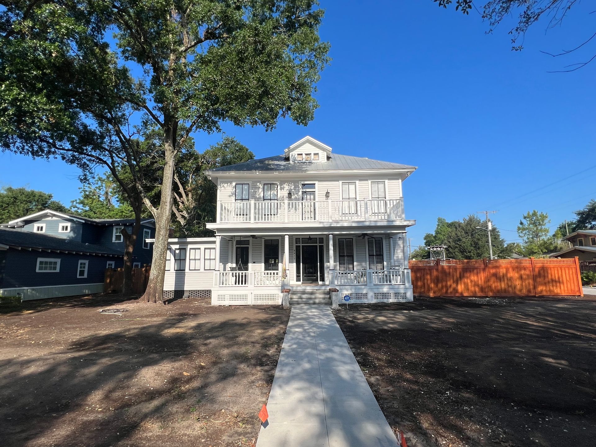 Two-story white house with porch and balcony, blue sky, concrete path, and bare earth.