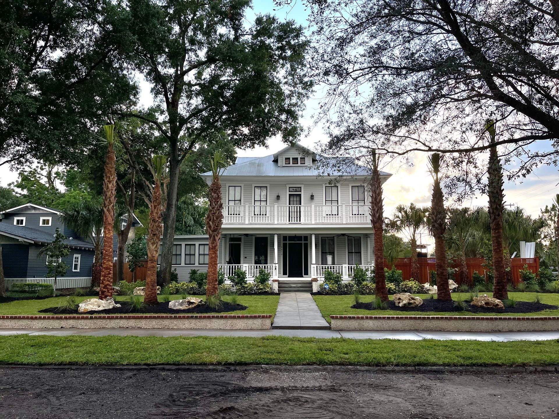 Two-story white house with front porch and palm trees on a tree-lined street.