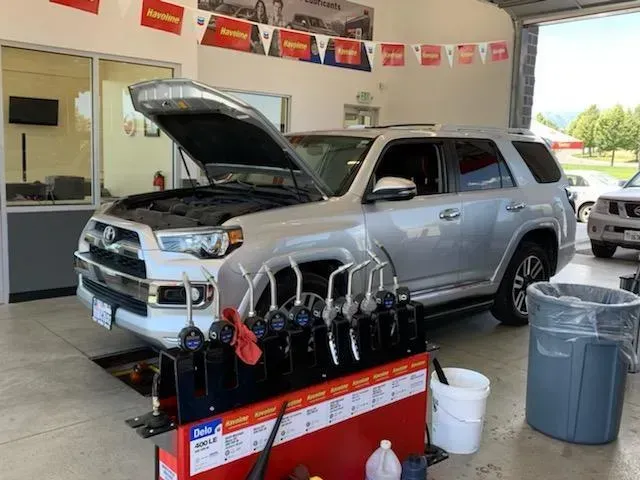 Silver SUV in a garage with hood open for service. Oil cans and equipment are nearby.