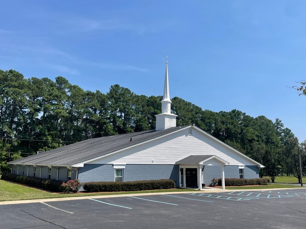 A church with a steeple is surrounded by trees and a parking lot.