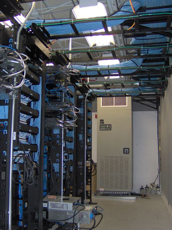 Server room with rows of black server racks, blue panels, and a large air conditioning unit.