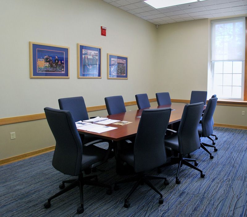 Meeting room with a long table, blue chairs, framed art, and a window with natural light.
