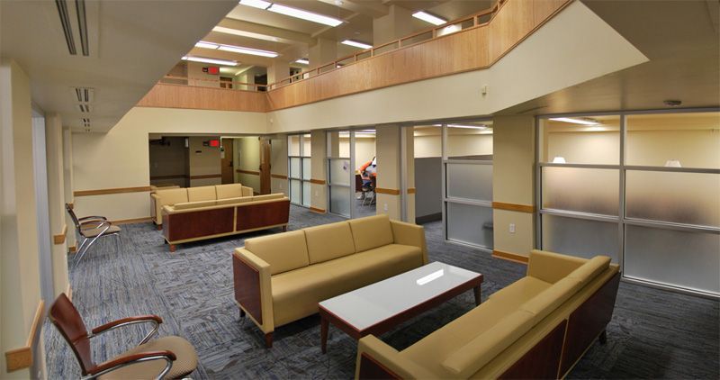 Lounge area with tan couches, white table, and carpeted floor; glass-walled offices visible.