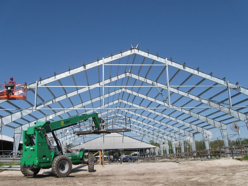 Construction of a large metal building under a bright blue sky; a green forklift is in the foreground.