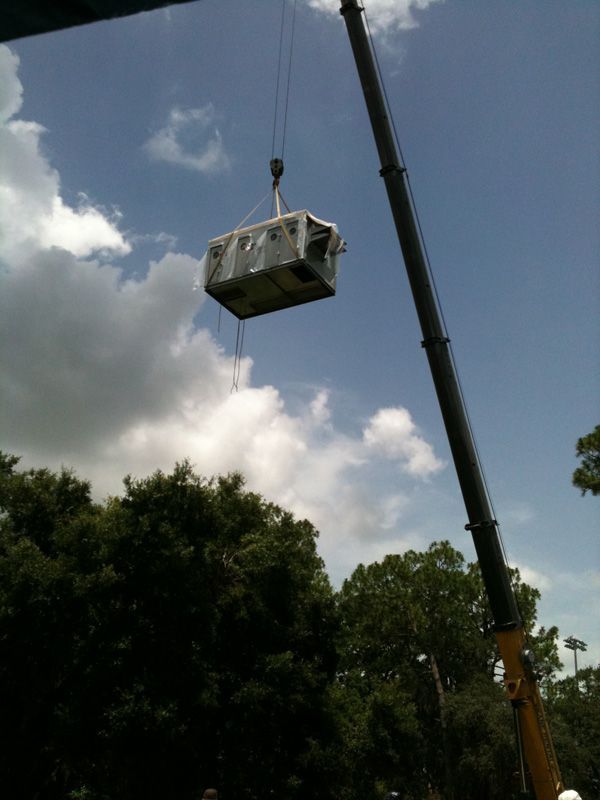Crane lifting a large, rectangular air conditioning unit against a cloudy sky.