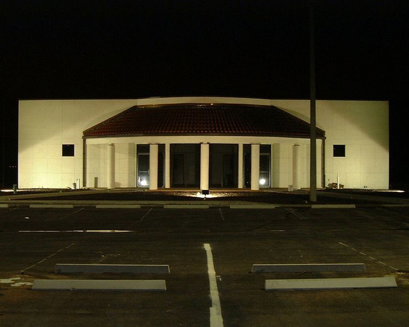 White building at night with a tile roof and columns over entrance, in an empty parking lot.