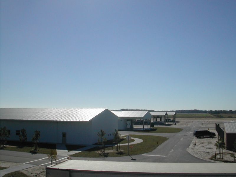 White industrial buildings with silver roofs under a clear blue sky. A paved road and sparse landscaping are visible.
