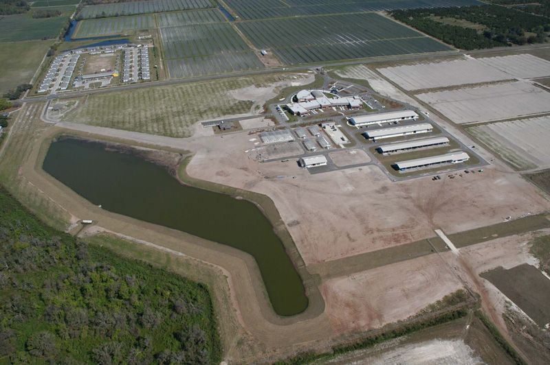 Aerial view of a farm complex with buildings, a pond, and fields. Green and brown tones dominate the landscape.