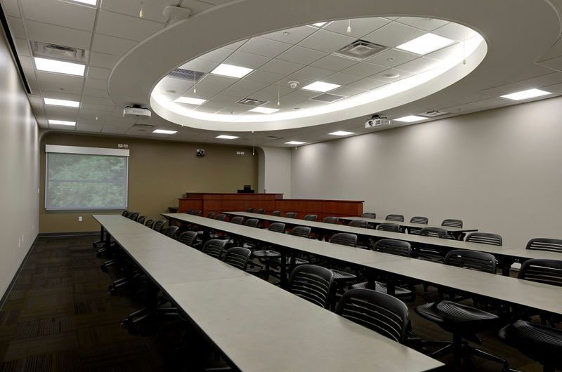 Empty classroom with long tables and chairs, projector screen, and recessed lighting.