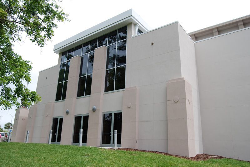 Beige building with large windows and glass doors, set against a green lawn and cloudy sky.