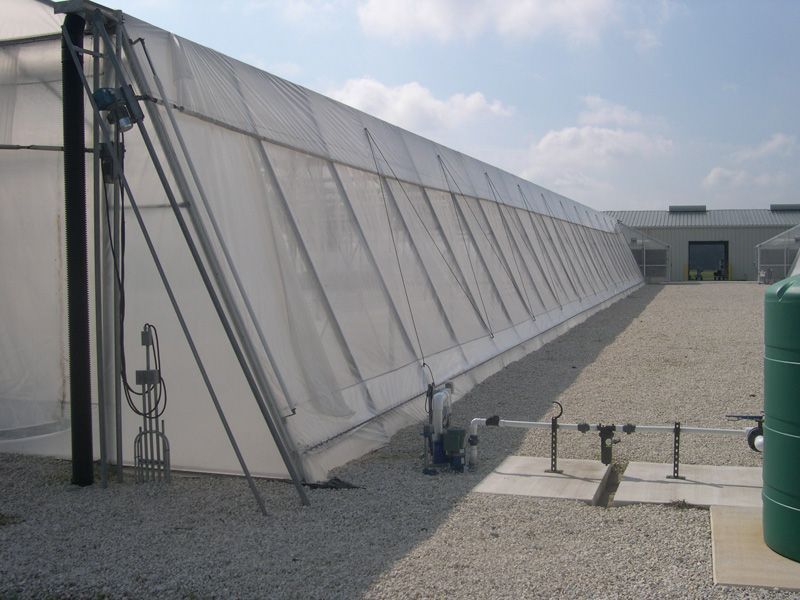 Long, white-walled greenhouse on gravel, with water pipes and a tall, black support pole.