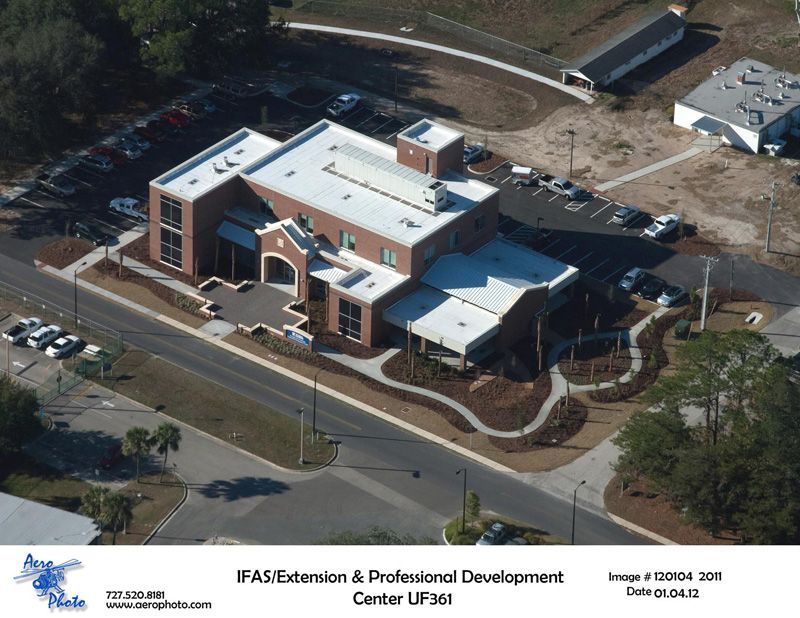 Aerial view of a brick building with a flat roof, surrounded by landscaping and parking, labeled “IFAS Extension & Professional Development Center”.