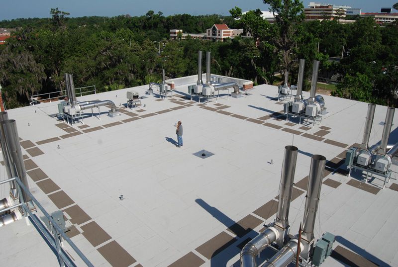 Man standing on a rooftop with multiple HVAC units and metal pipes. Trees and buildings in the background.