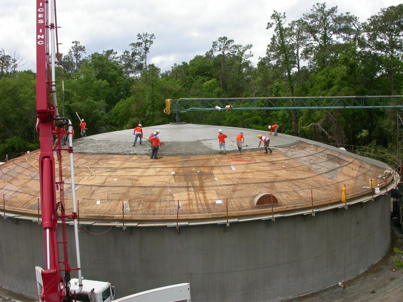 Construction crew pouring concrete onto a large, domed structure's roof. Red pump truck at the side.