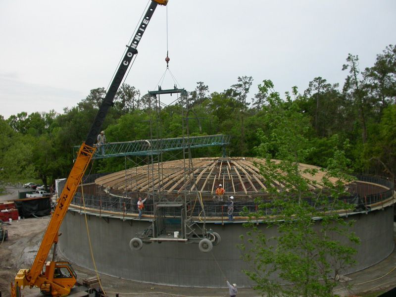 Crane lifting a metal structure onto a partially built circular concrete structure. Construction workers are present.