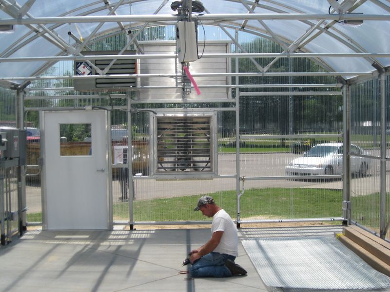 Inside a greenhouse, a man kneels working on the floor. Exterior visible through walls, a car and someone.