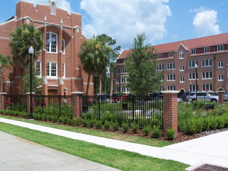 Brick buildings on a sunny day behind a black fence. Palm trees and green foliage in front.