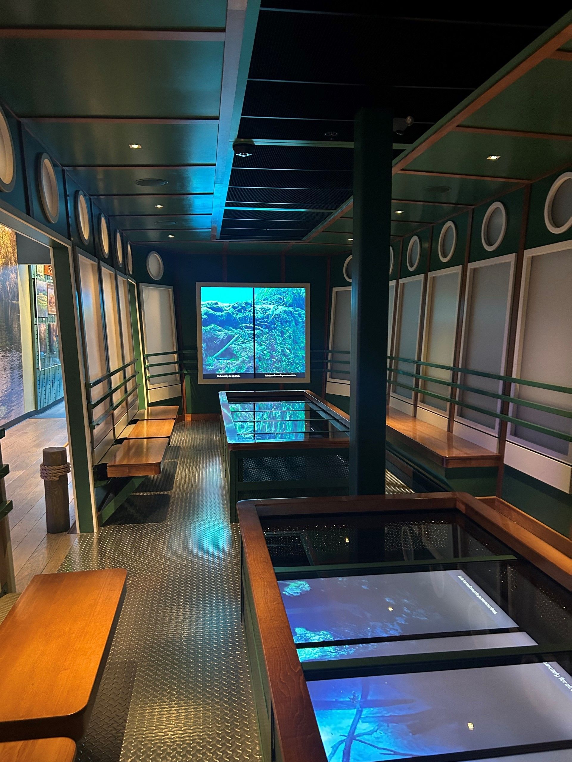 Restaurant interior with wood tables and benches, dark green walls, porthole windows, and a glass-bottomed water feature.