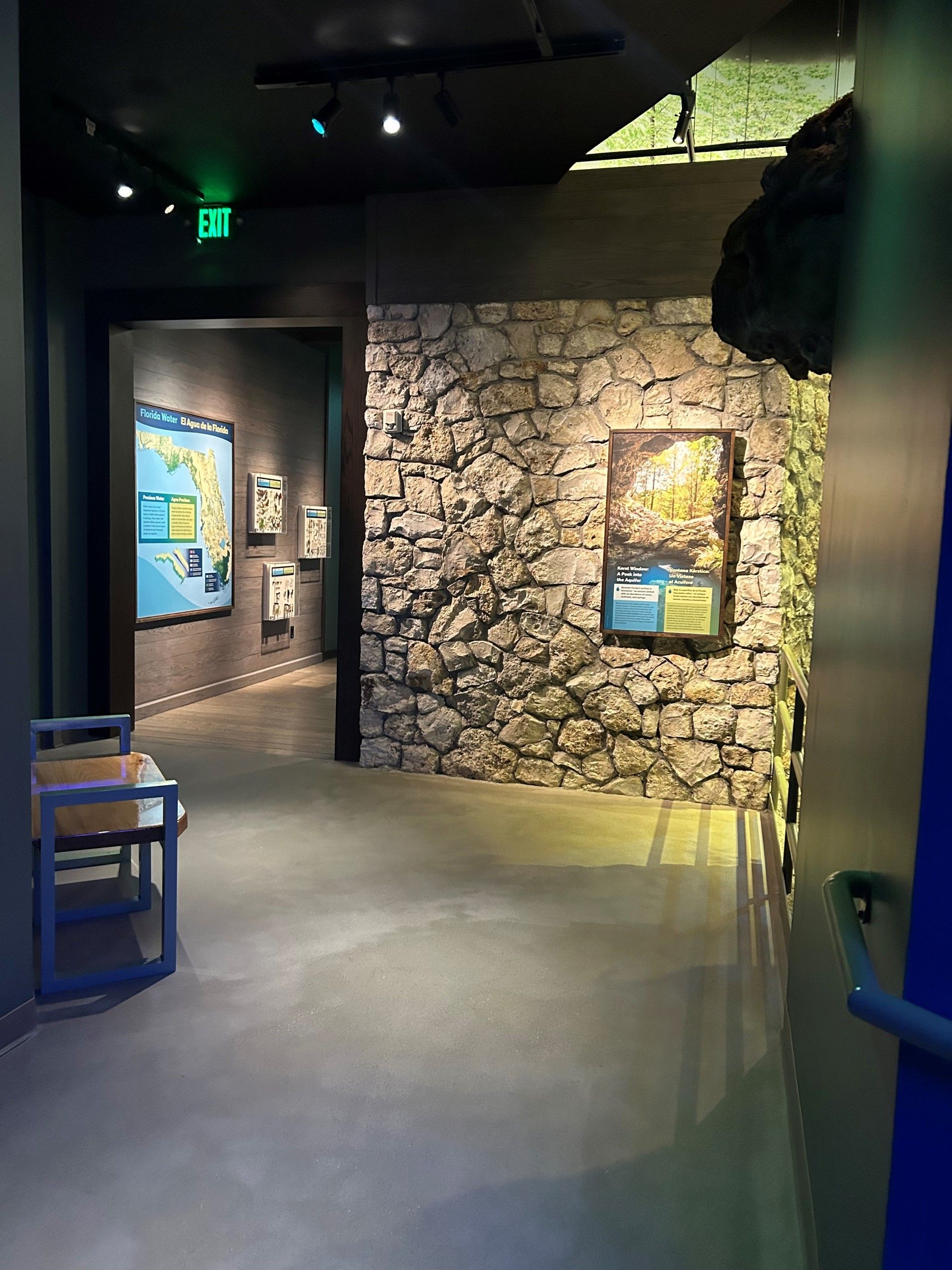 Hallway with stone wall, informational signs, and a blue chair. Dark colors, recessed lighting.