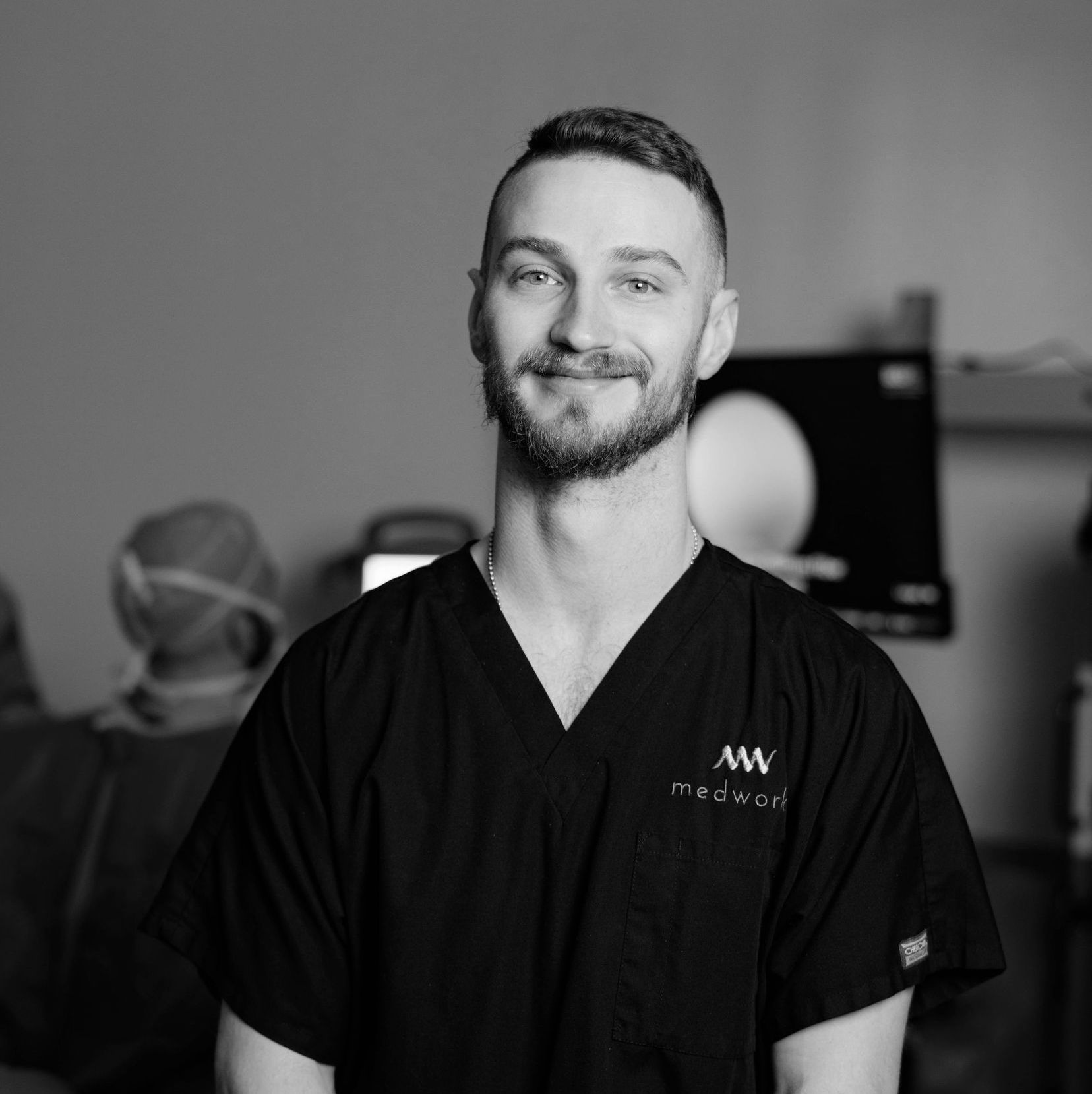 A man with a beard is wearing a black scrub top and smiling in a black and white photo.