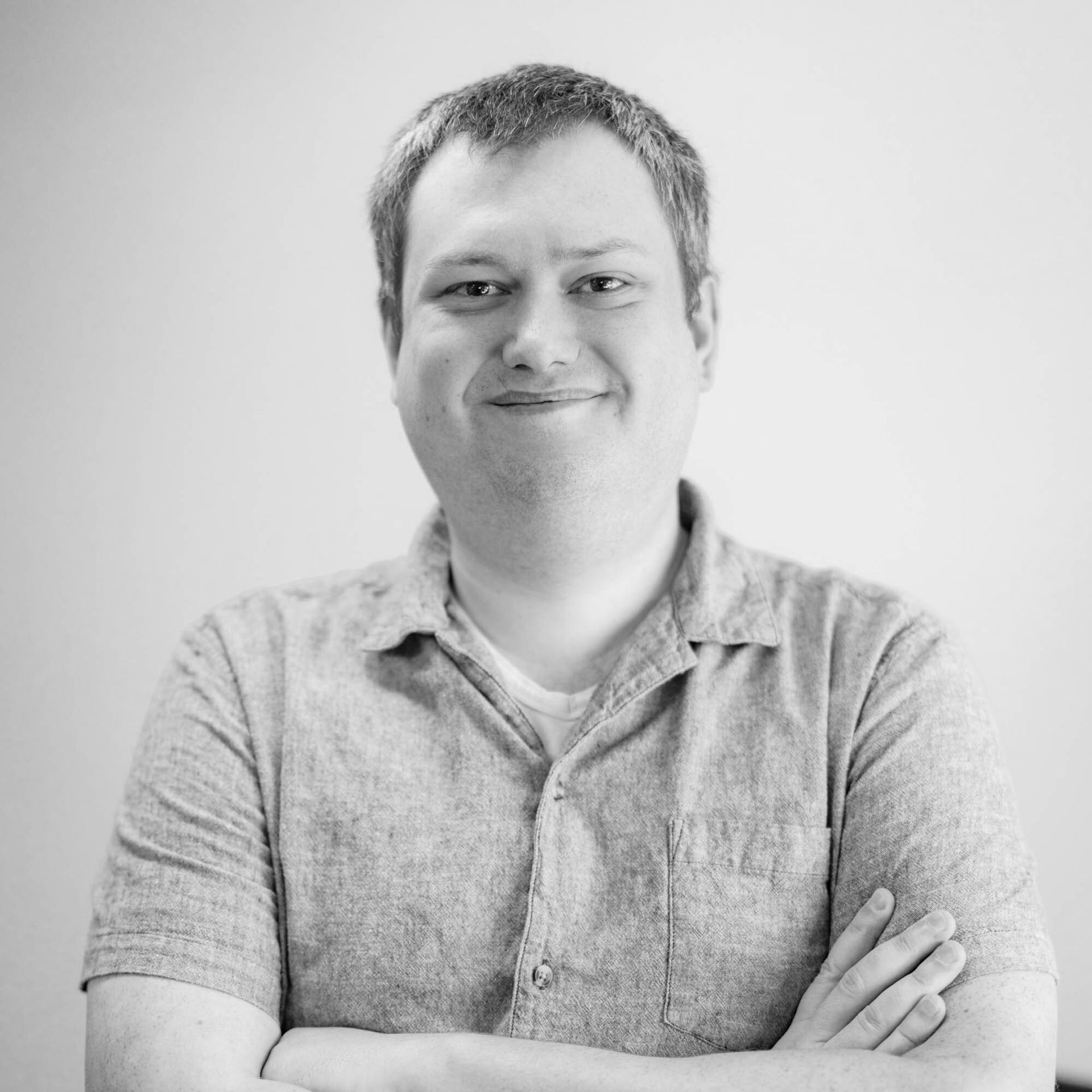 A man in a grey shirt is standing with his arms crossed and smiling.