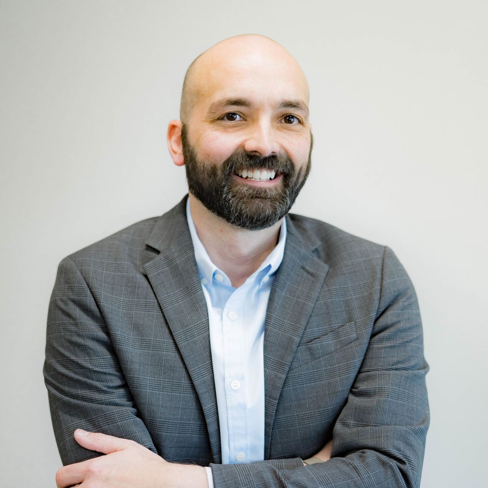 A bald man with a beard is wearing a suit and smiling with his arms crossed.