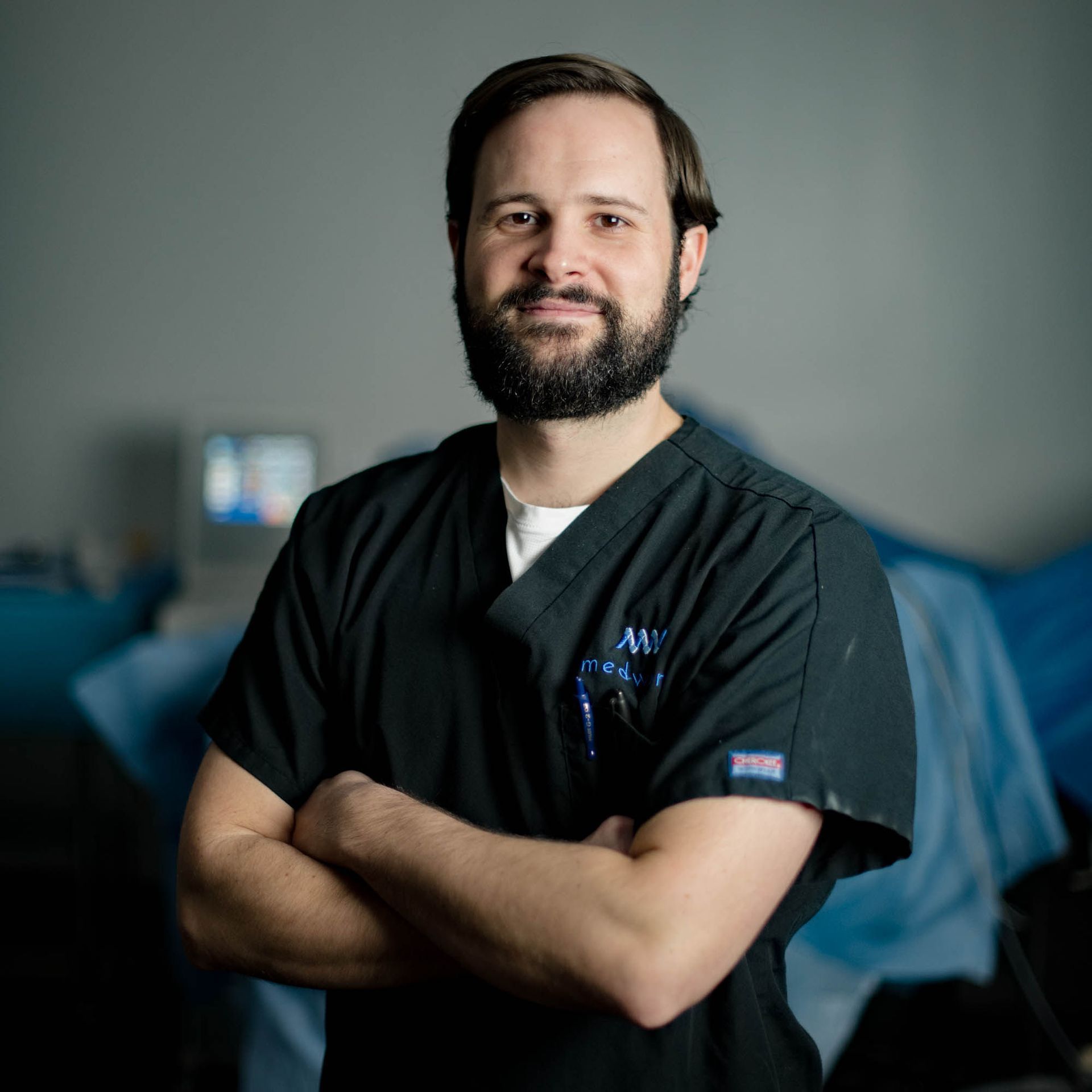 A man with a beard wearing a black scrub is standing with his arms crossed.