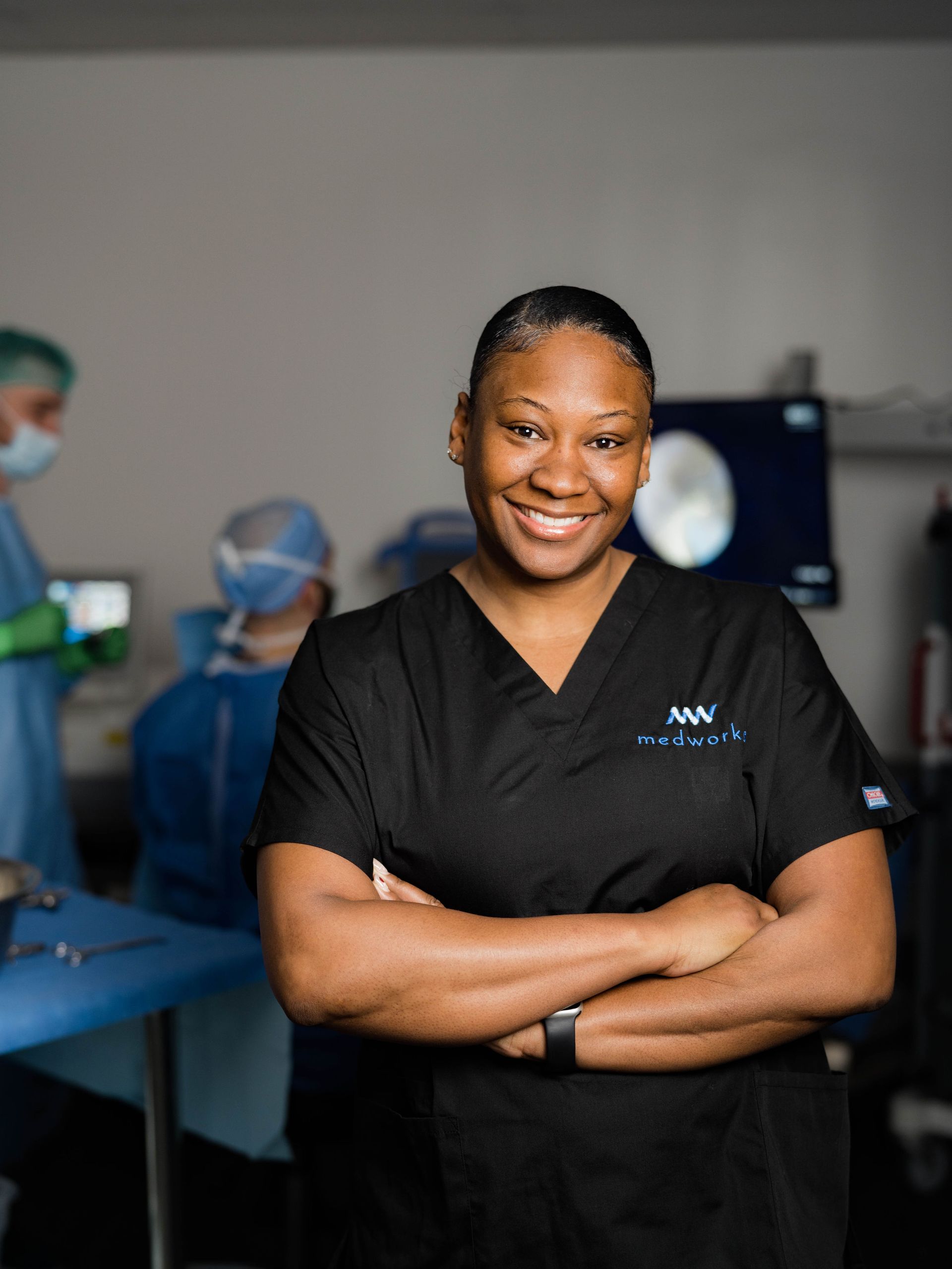 A woman in a black scrub is standing in an operating room with her arms crossed and smiling.