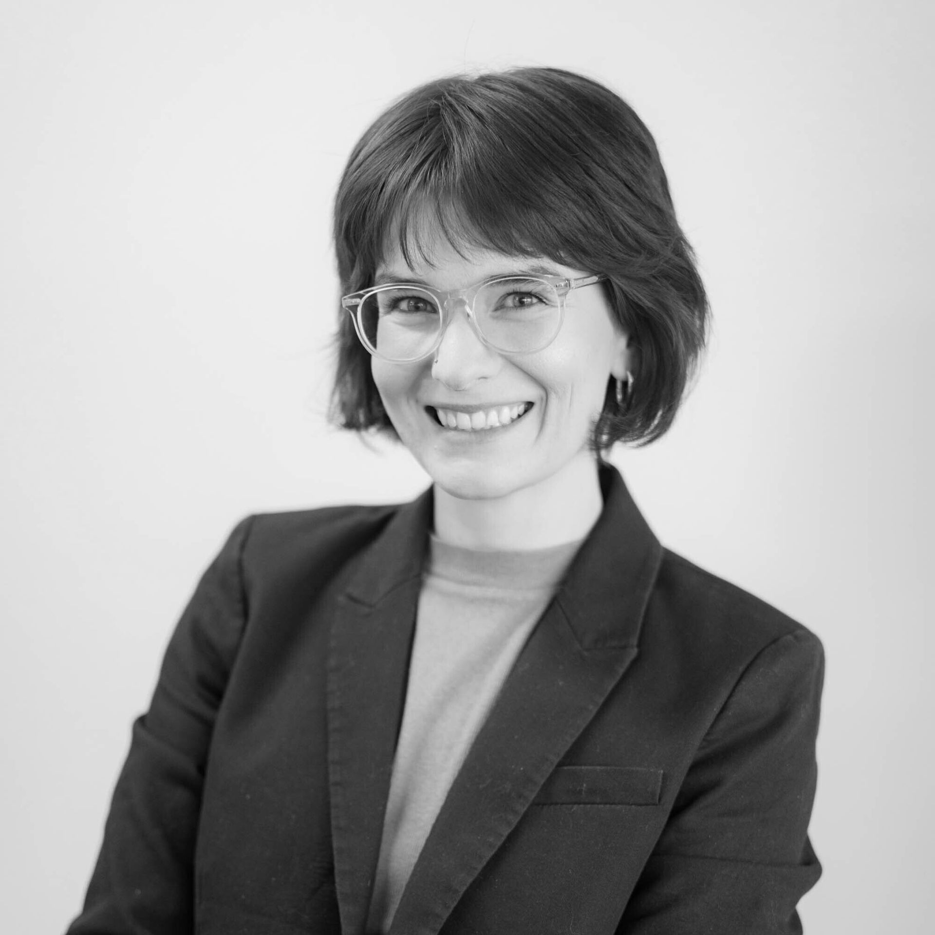 Smiling person in a dark blazer and glasses against a light background
