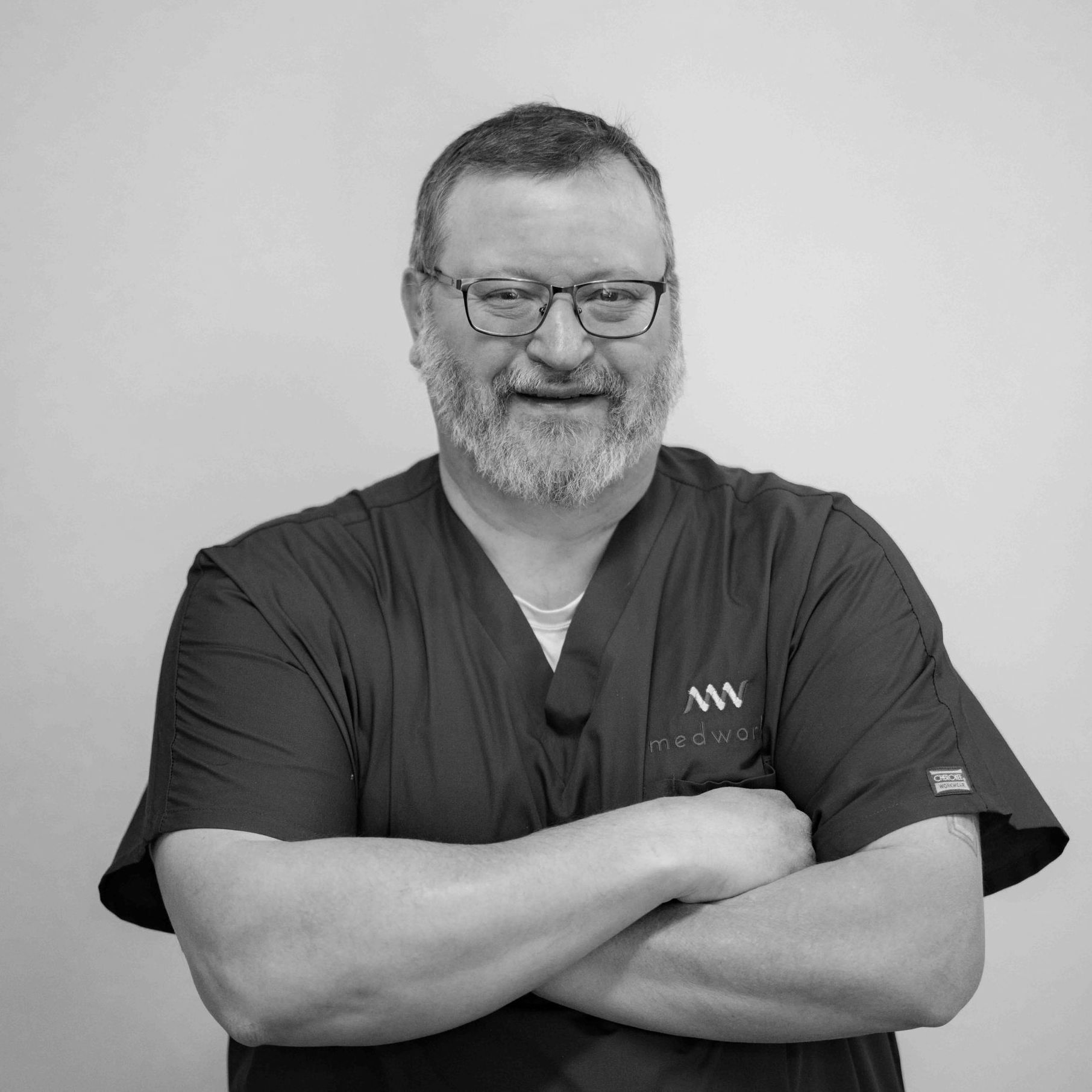 A man with a beard and glasses is standing with his arms crossed in a black and white photo.