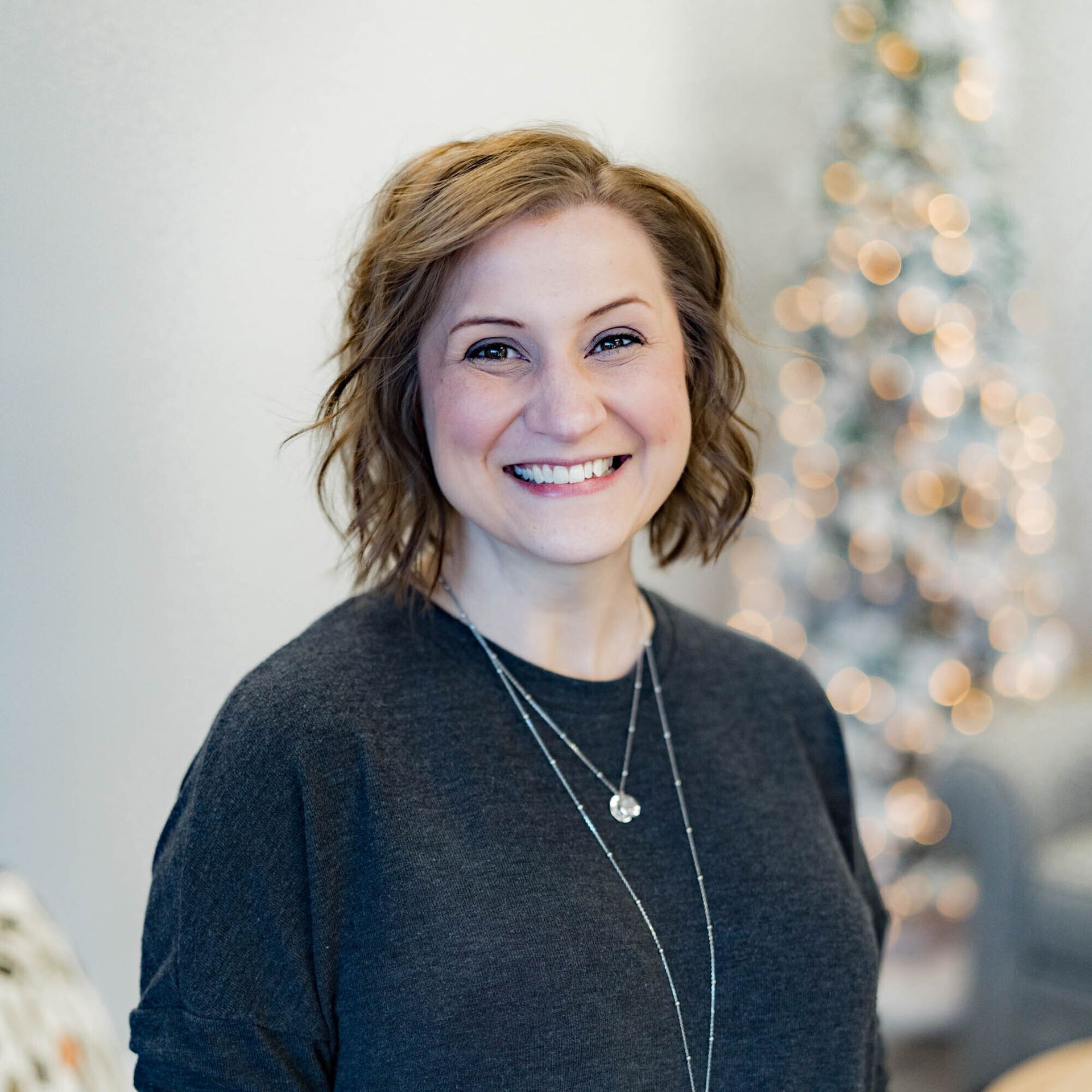 A woman wearing a black sweater and a necklace is smiling in front of a christmas tree.