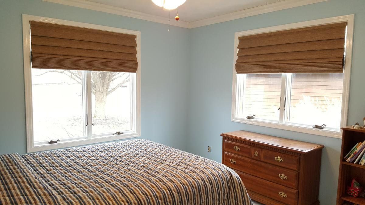 A bedroom with a bed , dresser , and two windows with roman shades.