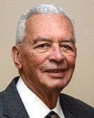 Studio headshot of a smiling man in a dark suit, white shirt, and tie against a beige background