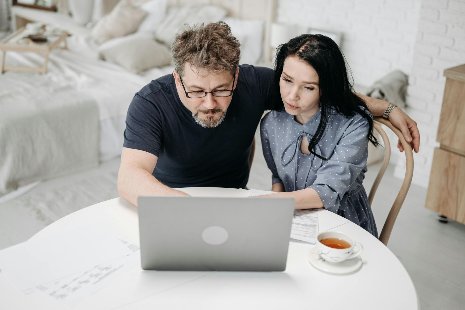 Two people lean over a laptop at a white table in a bright living room, with a cup of coffee nearby.