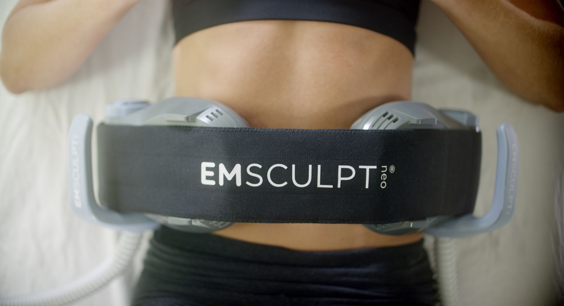 A woman is laying on a bed with a belt around her waist that says emsculpt.