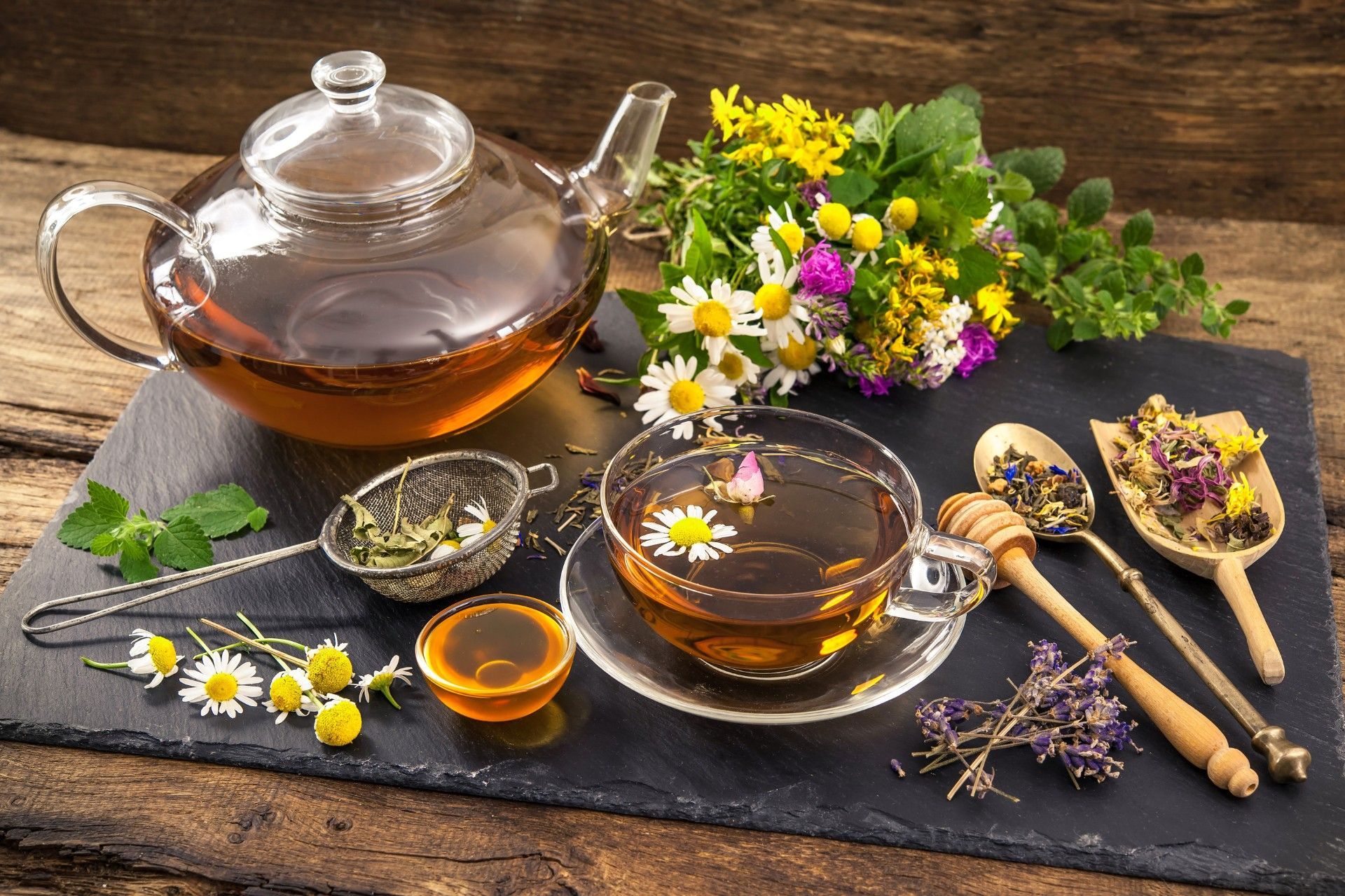 Glass teapot and cups of herbal tea with flowers, herbs, and honey on a rustic table