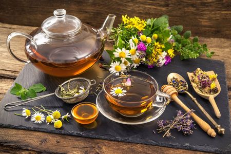 Glass teapot and cups of herbal tea with flowers, herbs, and honey on a rustic table