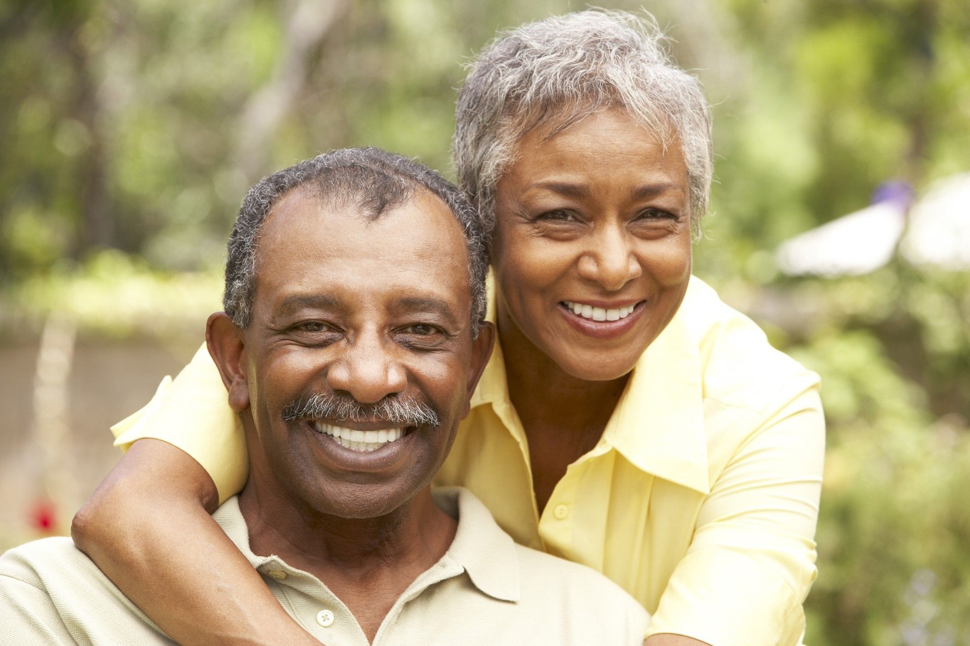 Smiling couple embracing outdoors in a sunny park, both wearing light-colored shirts.