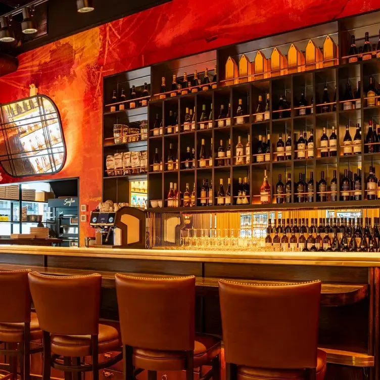 Bar interior with shelves of wine bottles, bar stools, and a mirror. Red wall behind the bar.