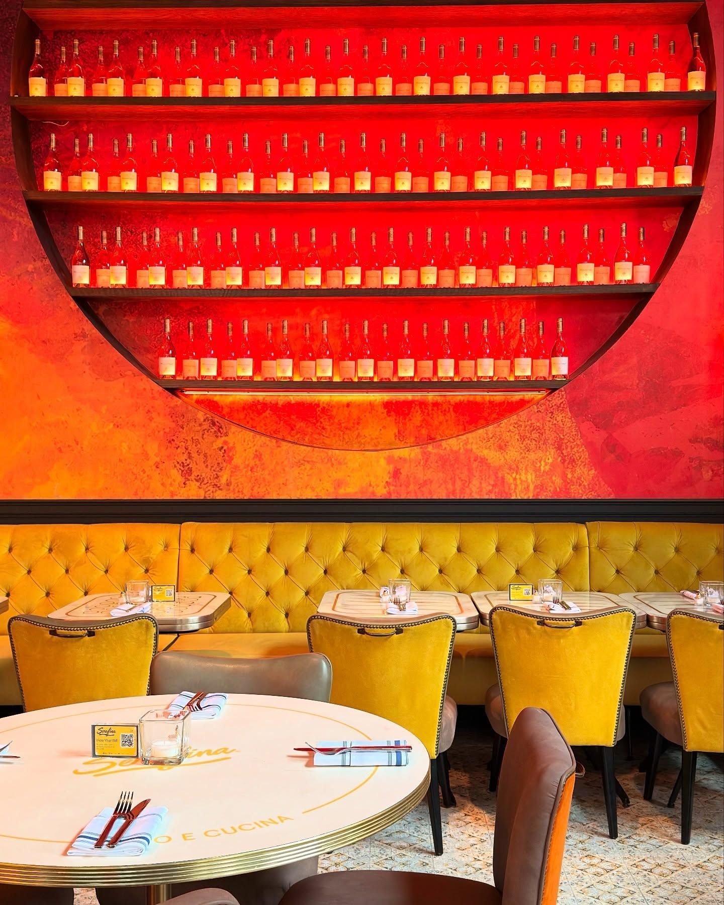 Restaurant interior with yellow booths, chairs, and a red-lit shelf of bottles.