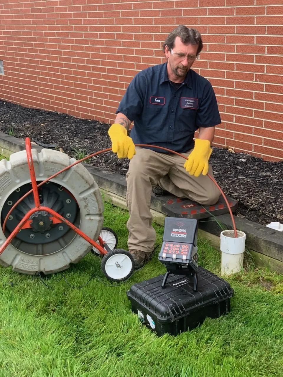 Plumber using a camera to inspect a pipe; he is wearing gloves, and the equipment is outside on grass.