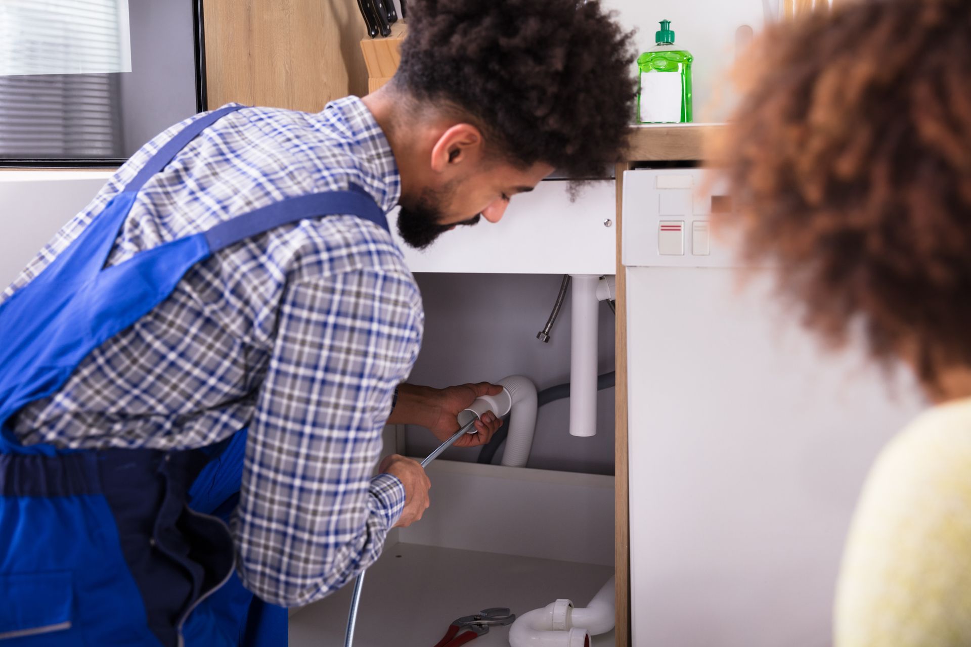 Plumber in blue overalls works under a kitchen sink while a person watches.